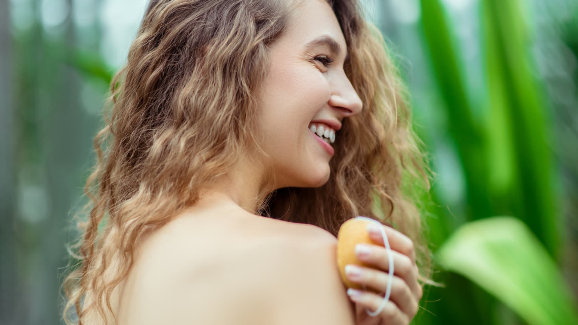 Blonde woman washing shoulder with plants in background.